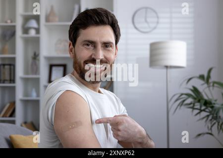 Ein lächelnder Mann zeigt stolz auf die Bandage am Oberarm nach der Impfung. Gesundheitsbegriff, moderne Medizin und persönliches Wohlbefinden. Entspannter und selbstbewusster Ausdruck, modernes Zuhause. Stockfoto