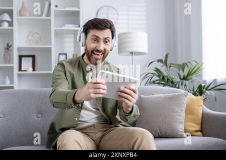 Aufgeregter Mann, der Kopfhörer trägt und Videospiele auf dem Tablet spielt, während er auf der Couch sitzt. Erleben Sie Freude und Unterhaltung während der Freizeit zu Hause. Stockfoto