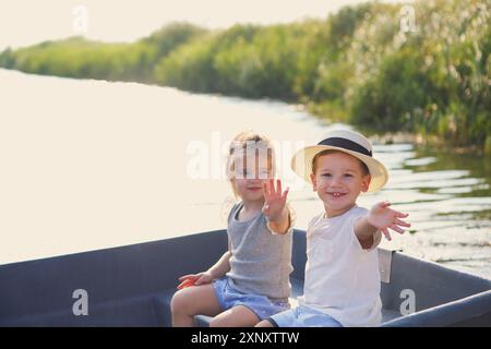 Fröhlicher Junge und Mädchen lächeln und winken die Hände, während sie im Boot in einem ruhigen Fluss auf dem Land sitzen Stockfoto