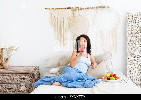 Glückliche junge Frau in der Erwartung, die roten Apfel hält und in die Kamera blickt, während sie auf dem Bett mit einem Teller Obst in einem hellen Boho-Stil mit sitzt Stockfoto