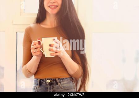 Junge Frau in legerer Kleidung sitzt auf Fensterbank und trinkt Tee, während sie zu Hause ruht Stockfoto