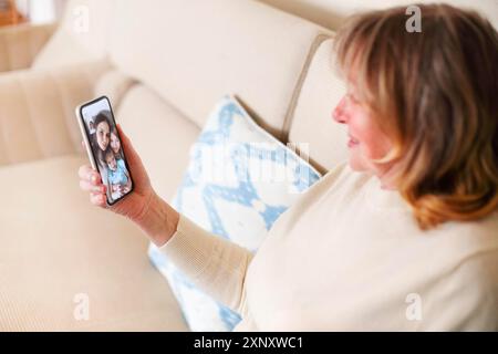 Hochwinkel-Seitenansicht einer fröhlichen Frau mittleren Alters, die auf dem Sofa sitzt und mit Tochter und Enkelkindern per Videochat auf dem Smartphone kommuniziert Stockfoto