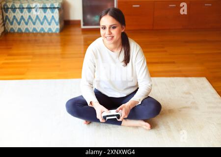 High Winkel der fröhlichen jungen Frau in Freizeitkleidung sitzen auf Teppich mit Konsole in den Händen und Videospiel spielen, während zu Hause ruhen Stockfoto