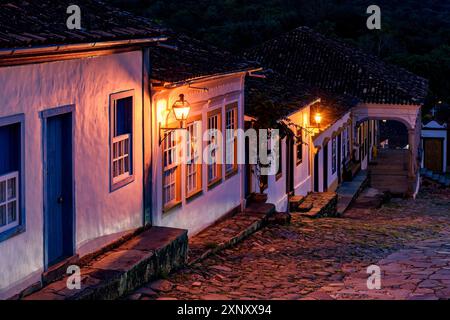 Blick auf eine alte gepflasterte Straße und die Häuser im Kolonialstil, die von Laternen in der historischen Stadt Tiradentes in Minas Gerais angezündet werden Stockfoto