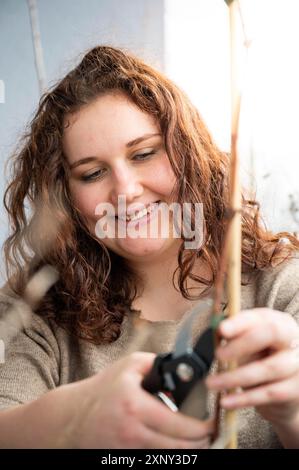 Nahaufnahme Frau mit braunem lockigem Haar, die in die Kamera schaut, während sie einen Baum auf ihrem Balkon schneidet, einen Ast mit Gartenscheren schneidet, ein zahniges Lächeln Stockfoto