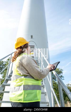 Ingenieurin mit braunem, lockigem Haar und gelbem Helm, die ihr digitales Tablet benutzt, während sie auf eine moderne Windenergieanlage mit erneuerbarer Energie blickt, Side Stockfoto