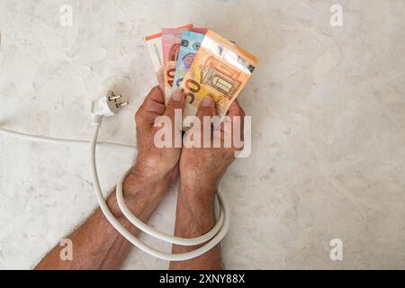 Die Hände eines Mannes, der Euro-Banknoten hält und mit einem Netzkabel mit Stecker verbunden wird, Konzept für steigende Stromkosten und Energieeinsparung Stockfoto