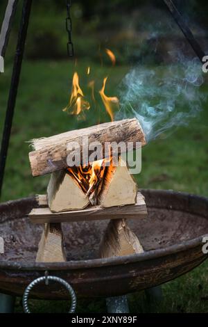 Geschickt angeordnete Holzscheite aus Brennholz, die mit Flammen und Rauch in einer Feuerschüssel unter einem Stativgrill in einem Garten für eine Grillparty brennen, Kopie Stockfoto