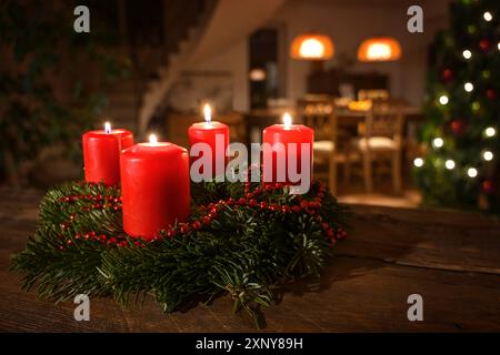 Dekorierter Adventskranz aus Tannenzweigen mit roten brennenden Kerzen auf einem dunklen Holztisch, Wohnzimmer mit Weihnachtsbaum im Hintergrund Stockfoto