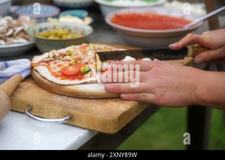 Hände schneiden frisch gebackene Pizza auf rustikalem Holzbrett, Zutaten im Hintergrund verschwimmen, Outdoor-Kochkonzept, ausgewählter Fokus, schmal Stockfoto