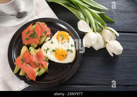 Spiegeleier in Herzform und andwiches mit Avocado und geräuchertem Lachs auf einem dunklen Teller, weiße Serviette und Tulpen, dunkelblauer Holztisch, Kopierraum Stockfoto