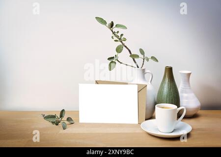 Modell einer leeren weißen Grußkarte mit einem naturbraunen Umschlag, drei Vasen mit einem Salbeizweig und einer Kaffeetasse auf einem Holztisch davor Stockfoto