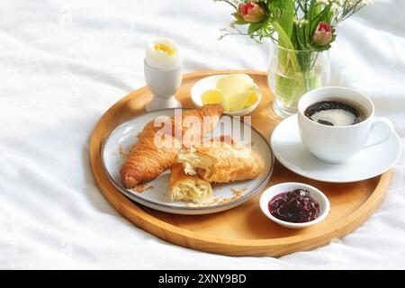 Kleines Frühstück mit Croissant, Kaffee, Marmelade und Ei auf einem Holztablett mit Blumenstrauß auf einem weißen überdachten Bett am Sonntag, Geburtstag, Valentinstag Stockfoto