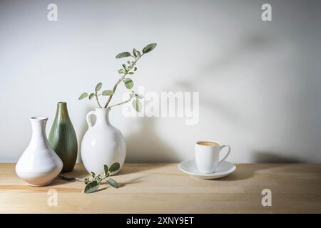 Helles Stillleben mit drei Vasen in weiß und grün, einem Salbeizweig mit Schatten und einer Kaffeetasse, Holztisch an einer grauen Wand, machen Sie eine Pause Stockfoto