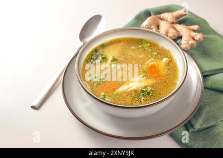 Hausgemachte Hühnersuppe mit Gemüse, Ingwer und Petersilie in einer Schüssel, grüne Serviette und Löffel auf einem schwulen weißen Tisch, Kopierraum, ausgewählter Fokus, schmal Stockfoto