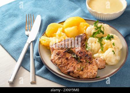 Gericht mit Schweinekotelett, Blumenkohl, Kartoffeln und Hollandaise-Sauce, serviert mit Kräutergarnitur auf einem Teller und einer blauen Serviette, ausgewählter Fokus, enge Tiefe Stockfoto