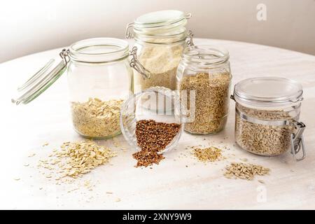 Getreide und Samen wie Haferflocken, Leinsamen, Sesam und Sonnenblumenkerne in Glasgläsern auf einem hellen Holztisch, gesunde Ernährung, Kopierraum Stockfoto