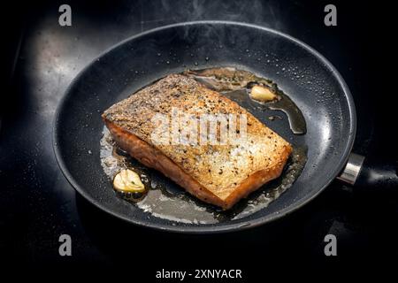 Frisches Lachsfilet mit knuspriger Haut wird mit Olivenöl, Kräutern und Knoblauch in einer schwarzen Pfanne auf dem Herd gebraten, Kopierraum, ausgewählter Fokus Stockfoto