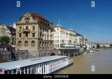 Elegante Gebäude am Fluss mit kleinen Kreuzfahrtschiffen unter hellblauem Himmel, Rhein, Basel, Schweiz Stockfoto