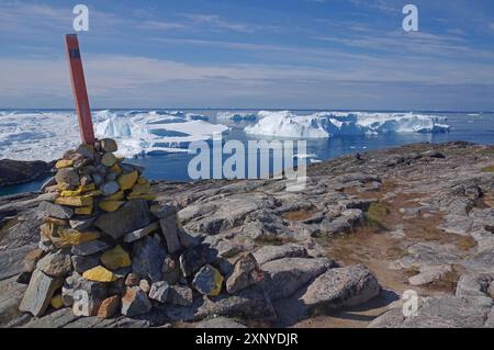 Ein Steinhaufen mit einer Flagge auf felsigem Gelände vor einer arktischen Landschaft und Eisbergen im Wasser, Wegweiser, Ilulissat, Disko Bay, Grönland Stockfoto