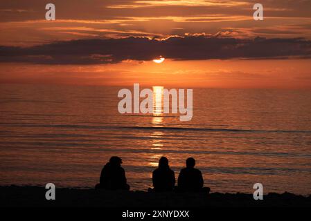 Drei Personen sitzen am Strand und beobachten den Sonnenuntergang über dem Meer, Silhouetten vor warmem Licht, Ostseehinsel Fischland-Darss-Zingst Stockfoto