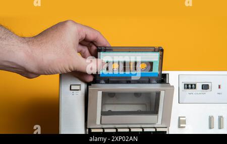 Vorderansicht der Hand des Mannes, die alte Musikkassette vor gelbem Hintergrund in den Alu-Recorder einsetzt Stockfoto