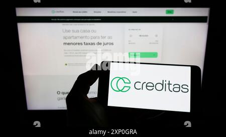 Person mit Handy mit Logo der brasilianischen Firma Creditas Solucoes Financeiras Ltda. Auf dem Bildschirm vor der Webseite. Schwerpunkt auf dem Display des Telefons Stockfoto