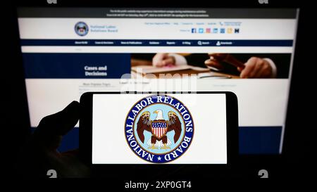 Person, die ein Mobiltelefon hält, mit dem Logo des US National Labor Relations Board (NLRB) auf dem Bildschirm vor der Webseite. Schwerpunkt auf dem Display des Telefons Stockfoto