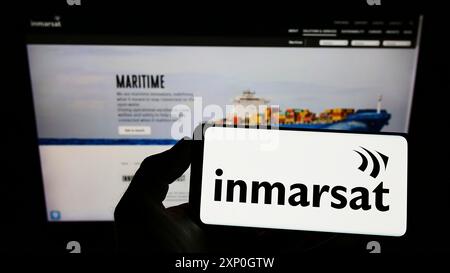 Person, die ein Mobiltelefon mit dem Logo des Satellitenunternehmens Inmarsat Global Limited auf dem Bildschirm vor der Unternehmenswebseite hält. Schwerpunkt auf dem Display des Telefons Stockfoto