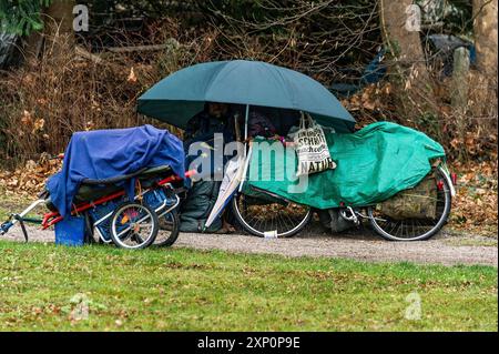 Obdachloser Mann im Lager auf Parkbank unter Schirm mit Decke und Schlafsack, Fahrrad mit Plane bedeckt, Fahrradanhänger mit Personal Stockfoto