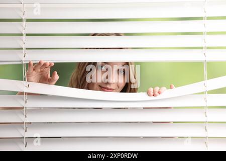 Junge Frau, die durch Fensterläden auf verschwommenem Hintergrund schaut Stockfoto