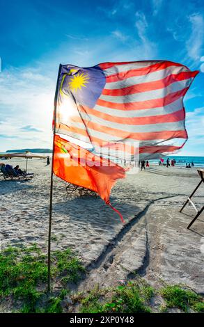 Am Strand von Cenang, der in den Sand gepflanzt ist, schlägt die malaysische Flagge in den Winden, die aus der Andamanensee kommen, wenn die Leute hinter dem Gleitsegel laufen und entlang laufen Stockfoto