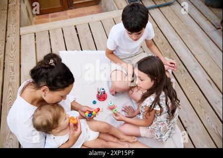 Mutter und drei Kinder genießen im Sommer Spielzeit mit Spielzeug im Freien auf einer Holzterrasse. Familienbindung, Geschwisterinteraktion und Kinderfreude. Stockfoto