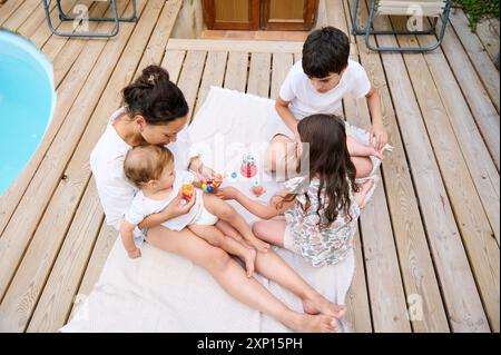 Eine Mutter genießt mit ihren drei Kindern einen schönen Moment auf einer Holzterrasse am Pool, spielt mit farbenfrohen Spielzeugen und zeigt die Familie Stockfoto