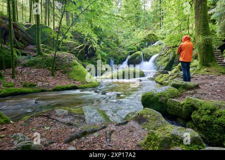 Eine Person in einer orangefarbenen Jacke steht an einem ruhigen Wasserfall, umgeben von üppigem Grün und moosbedeckten Felsen, die die Gelassenheit verkörpern. Europa, Deutschland, Stockfoto