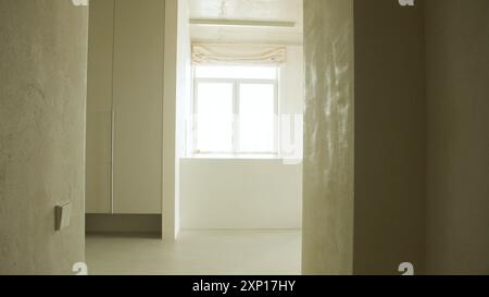Das Innere des leeren Wohnzimmers mit großem hellen Fenster und Vorhängen durch eine geschlossene Tür in einem modernen Apartment mit weißen Wänden und Hartholzboden Stockfoto