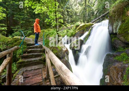 Ein großer Mann in einer orangefarbenen Jacke steht auf einer Holzbrücke und beobachtet einen Wasserfall, der von üppigem Grün umgeben ist. Europa, Deutschland, Schwarzwald, Bühlertal, Stockfoto