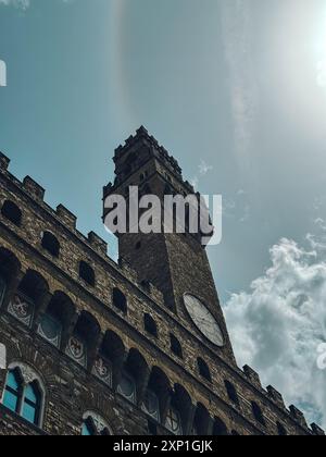 Ein hoher Turm mit einer Uhr drauf. Der Himmel ist bewölkt und die Sonne scheint. Der Turm ist von Gebäuden umgeben Stockfoto