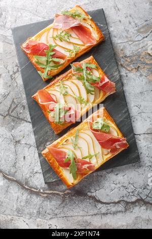 Köstliche gebackene Blätterteiggebäck mit Birnen, Frischkäse, Marmelade und Rucola aus nächster Nähe auf einem Marmorbrett auf dem Tisch. Vertikale Draufsicht von oben Stockfoto