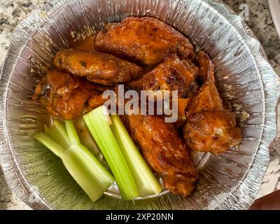 Eine köstliche Portion scharfer Hähnchenflügel wird mit frischen Selleriestangen kombiniert, die in einem glänzenden Folienbehälter präsentiert werden. Die Flügel sind perfekt gekocht, s Stockfoto