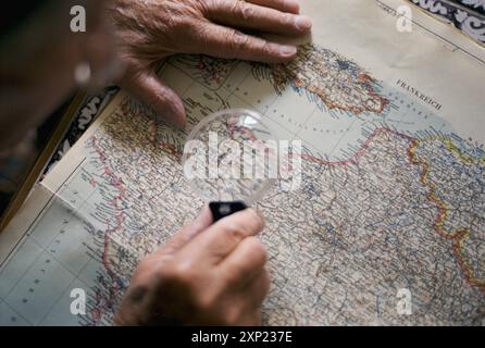 Nahaufnahme der Hände mit einer Lupe, um eine detaillierte historische Karte von Frankreich zu betrachten, die die Normandie hervorhebt. Eine Metapher für die Erforschung von Geschichte und Erinnerungen. Stockfoto