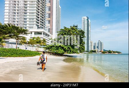 Eine Dame, die am Tanjing Tokong Strand in Penang, Malaysia, mit hohen, modernen Wolkenkratzern spaziert, Teil einer Skyline der Stadt im Hintergrund. Stockfoto