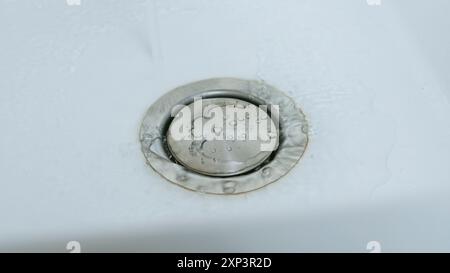 Nahaufnahme eines Edelstahlabflusses in einem weißen Waschbecken. Wasser fließt in Richtung Abfluss. Stockfoto