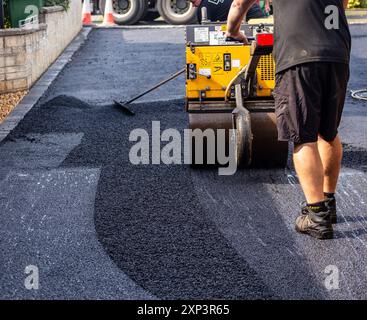 Arbeiter, der an sonnigen Tagen eine Asphaltwalzenmaschine bedient, während er eine neue Fahrbahnoberfläche pflastert. Stockfoto