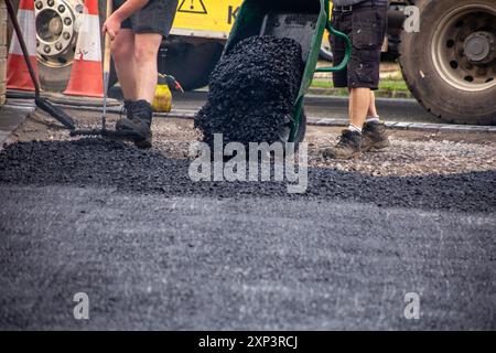 Arbeiter legen neuen Asphalt auf einer neuen Einfahrt-Baustelle ab, während einer aus einer Schubkarre gießt und der andere den Asphalt mit einem Rechen ebnet. Stockfoto