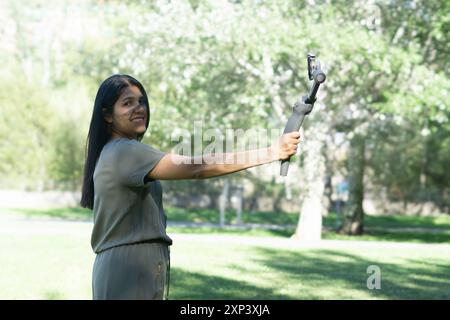 Eine Frau hält eine Kamera und lächelt. Die Kamera ist ein kleines schwarzes und silbernes Gerät. Die Frau steht in einem Park, umgeben von Bäumen und Gras Stockfoto