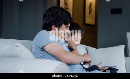 Ein Junge und ein Erwachsener sitzen auf einem Sofa und konzentrieren sich gemeinsam auf ein Tablet. Moment der Verbindung und Interaktion mit moderner Technologie, die Familienleben hervorheben Stockfoto