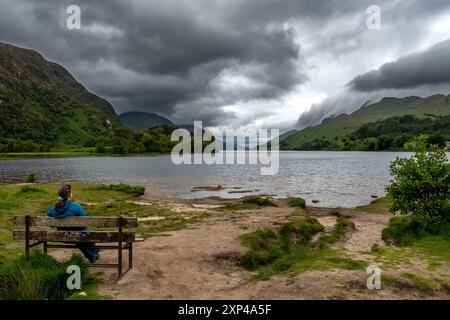 Frau auf der Bank, die über den Lake Loch Shiel am Glenfinnan Monument on the Road to the Isles in Lochaber in Schottland, Großbritannien, blickt Stockfoto