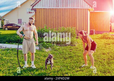 Ein amerikanischer Tyrannehund trinkt Wasser aus dem Gartenschlauch im Hinterhof des Landhauses und unterhält seine jungen Besitzer, Mann und Frau in ihren 20ern Stockfoto