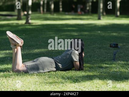 Eine Frau liegt auf dem Gras und macht ein Foto mit einer Kamera. Die Szene ist friedlich und entspannend, die Frau genießt die Natur und fängt ein Stockfoto